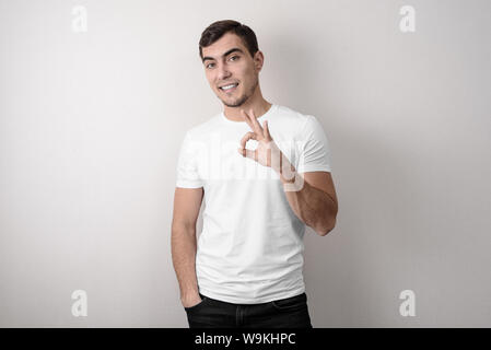 Junge europäische Mann im weißen T-Shirt auf grauem Hintergrund zeigt OK Zeichen, Studio Aufnahmen Stockfoto