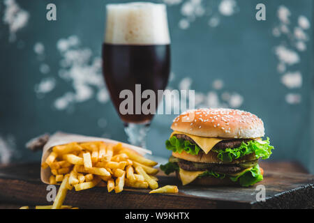 Burger mit Kartoffeln und dunkles Bier auf einem Holzbrett auf ein blau-grauer Hintergrund Stockfoto