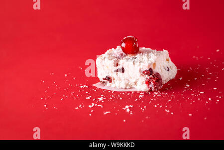 Stück White Christmas Cake mit Kokosnuss, getrocknete Früchte, Kirschen, auf einem roten Hintergrund. Australische traditionelle süße Speisen. Winterurlaub Dessert Stockfoto