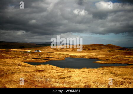 Dunkle und launische irische Landschaft von Connemara, einsames weißes Haus inmitten von goldenem Gras, umgeben von dunklen Wolken und hohen Bergen Stockfoto