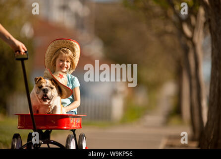 Mädchen und ihre Bulldogge, beide tragen Cowboyhüte, in einem roten Wagen gezogen. Stockfoto