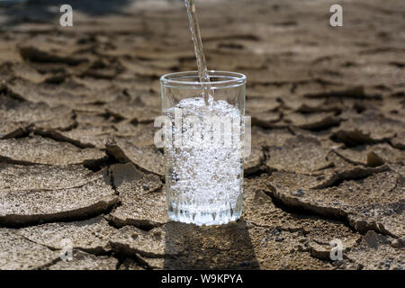 Reine klare, frische Wasser wird in einem Becherglas steht in der Mitte einer trockenen Risse im Lehm wüste Land gegossen Stockfoto