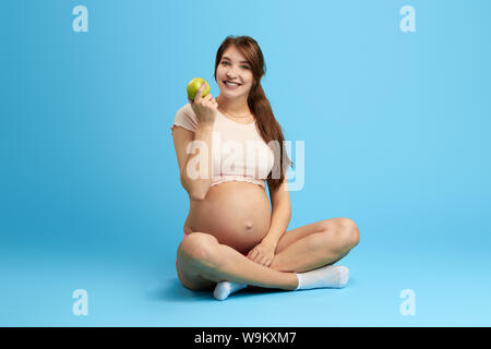 Junge glücklich charmante schwangere Frau mit Apple auf dem Fußboden sitzen. gesunde organische Ernährung. in voller Länge Foto. Isolierte blauen Hintergrund. gesund Stockfoto