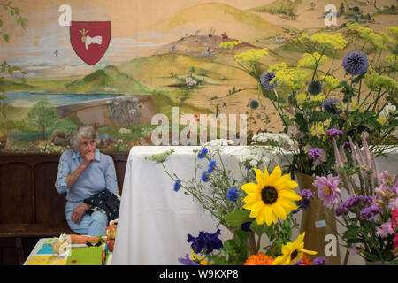 Allein alt werden, singen Sie ältere Frauen bei der Dorfproduktion und Blumenausstellung, allein im Rathaus, wo die Ausstellungen bewertet werden. Das Wandgemälde an der Dorfhalle ist von ihrer Gemeinde in den Brendon Hills, Exmoor. Das Schild des Ralph-Familienwappens. Brompton Ralph, Somerset, The West Country, England 10. August 2019 HOMER SYKES Stockfoto