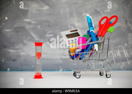 Warenkorb für Schulbedarf und Sanduhr auf grauem Hintergrund. Shopping Zeit zur Schule Stockfoto
