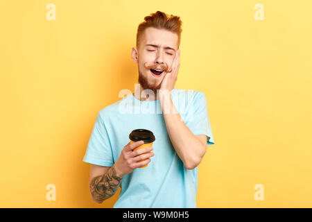 Verschlafene mann Gähnen als schlechte Nacht, trinken Kaffee erfrischt fühlen, trägt das stilvolle blaues T-Shirt, über gelbe Wand isoliert. Müdigkeit Konzept. Stockfoto