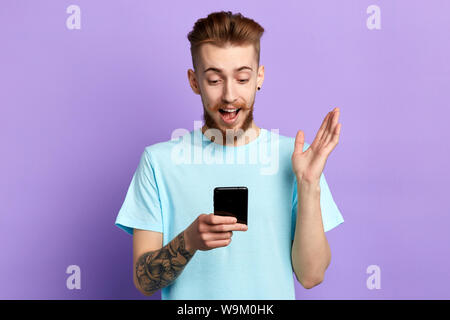 Lächelnden jungen stilvollen Mann auf seine Smart Phone während Text auf blauem Hintergrund isoliert Messaging. Mann an guten Nachrichten, die er Fr gelesen hat Freude Stockfoto