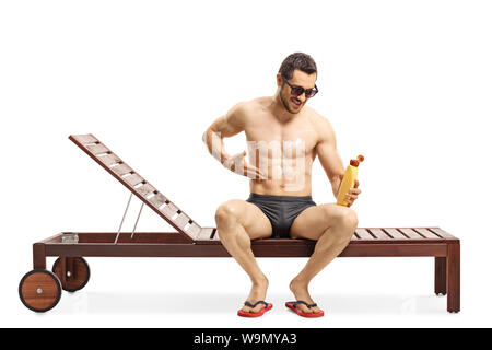 Volle Länge geschossen von einem jungen Mann der Anwendung Sun Creme und sitzen auf einer Sonnenliege auf weißem Hintergrund Stockfoto