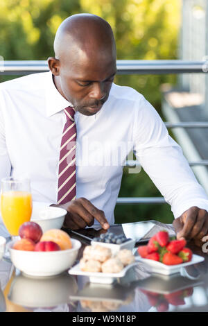 Afrikanischer Mann das Frühstück draußen auf der Terrasse essen, in der Kleidung, auf einem Tablet-PC oder das ipad arbeiten Stockfoto