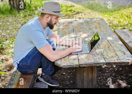 Junge hübsche kaukasischen bärtigen Mann mit Hut auf dem Kopf sitzt auf dem Holztisch in Wald und Arbeiten mit dem Computer. Stockfoto