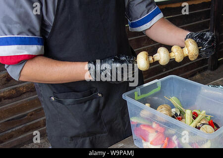 Сhef Köche Pilze. Mann in Uniform und Handschuhe vorbereiten champignon Spiesse zum Grillen. Außerhalb Picknick party Küche mit vegetarischen Essen Stockfoto