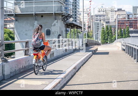 Ein junges Mädchen mit ihrem Haar und Rucksack Fahrten eine vermietet orange Bike über die Brücke und gleichzeitig fließt am Telefon, die den Lebensstil Stockfoto