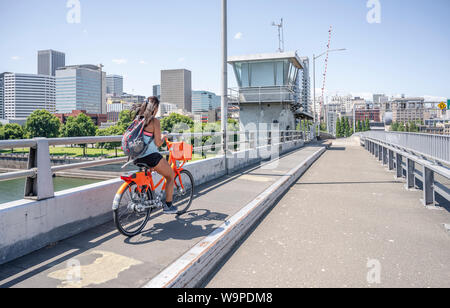 Ein junges Mädchen mit ihrem Haar und Rucksack Fahrten eine vermietet orange Bike über die Brücke und gleichzeitig fließt am Telefon, die den Lebensstil Stockfoto