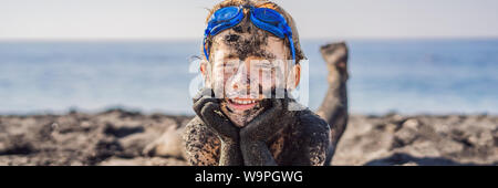 BANNER, LANGE FORMAT Schwarzer Freitag Konzept. Lächelnde Junge mit schmutzigen schwarzen Gesicht sitzen und spielen auf schwarzem Sand Meer Strand vor dem Schwimmen im Ozean Stockfoto