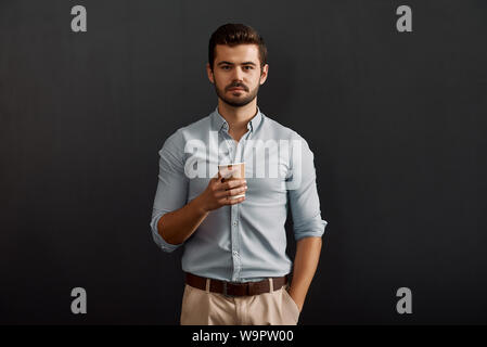 Ich brauche eine Pause. Junge bärtige Mann hält eine Tasse heißen Kaffee und Suchen an der Kamera, während gegen den dunklen Hintergrund stehen. Kaffeepause. Studio shot Stockfoto