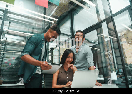 Glücklich zusammen zu arbeiten. Gruppe von drei Jungen und positive Mitarbeiter mit modernen Technologien und lächelnd, während im modernen Büro. Job Konzept. Arbeitsplatz Stockfoto