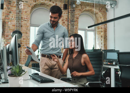 Wir haben tolle Ergebnisse jungen gutaussehenden Mann in legere Kleidung, etwas, das seine digitale Tafel zu seiner schönen weiblichen Mitarbeiter in modernen Büro. Arbeitsplatz. Geschäftskonzept. Stockfoto