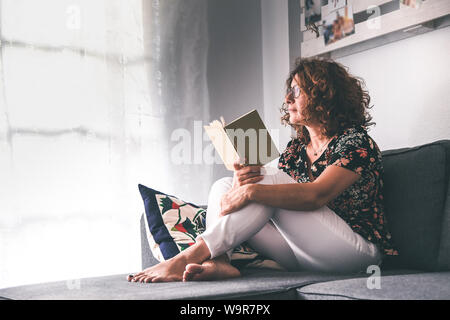 Schöne Mädchen mit einem Buch auf dem Sofa zu Hause sitzen. Frau mittleren Alters entspannen und genießen Freizeit freie Zeit außerhalb des Fensters. Nachdenklich Stockfoto