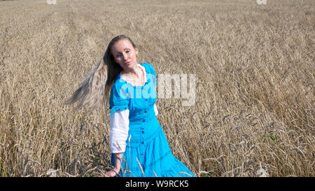 Schlanke junge Frau in einem blauen Kleid mit langen blonden Haare im Wind flattern in einem Feld der goldene Weizen Ohren im Sommer Stockfoto