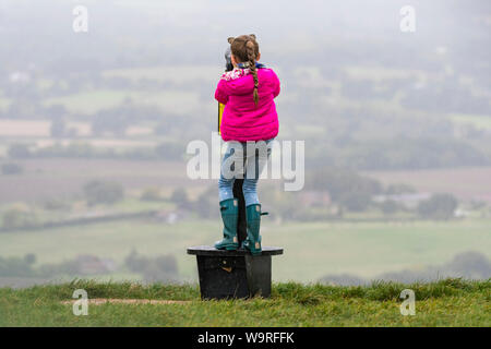 Junges Mädchen mit einem öffentlichen Teleskop an der Aussicht auf der Suche von einem Hügel in der Britischen Landschaft an einem regnerischen Tag in East Sussex, England, UK. Stockfoto