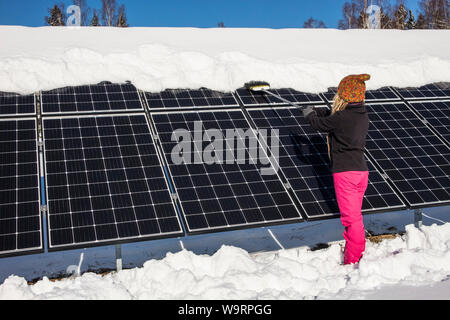 Frau schieben Schnee Solarmodule aus im Winter. Wenn Schnee Panels umfasst, können Sie nicht Strom erzeugen. Die kleinen privaten Home Haus im Hintergrund. Stockfoto