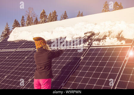 Frau schieben Schnee Solarmodule aus im Winter. Wenn Schnee Panels umfasst, können Sie nicht Strom erzeugen. Die kleinen privaten Home Haus im Hintergrund. Stockfoto