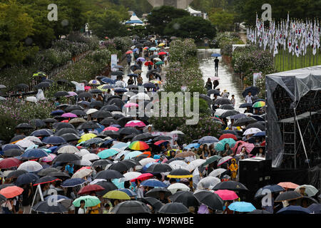Cheonan, Südkorea. 15 Aug, 2019. 15.August 2019 - Cheonan, South Korea-People holding Sonnenschirme sehen Sie hier einen TV-Bildschirm während der Veranstaltung für den 74. Jahrestag der Unabhängigkeit Koreas Tag an der Halle der Unabhängigkeit Koreas in Cheonan, Südkorea, am Donnerstag, 15 August, 2019. Quelle: Bloomberg über Pool/ZUMA Draht/Alamy leben Nachrichten Stockfoto