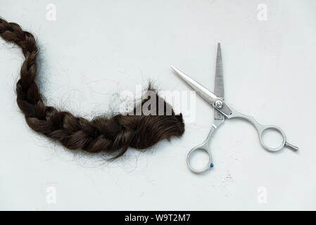 Friseur Schere mit geschnittenen pigtail von braunes Haar auf grau Holz- Hintergrund. Kopieren Sie Platz. Ansicht von oben. Flach Stockfoto