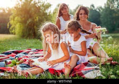 Mutter, Großmutter und Kinder weben Flechten, während die jüngsten Buch liest. Familie Spaß beim Picknick im Park. Drei denerations Stockfoto
