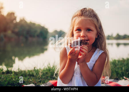 Kleines Mädchen Kuchen essen bei Familie Picknick im Sommer River. Kind holding Stück Kuchen und lächelnd Stockfoto