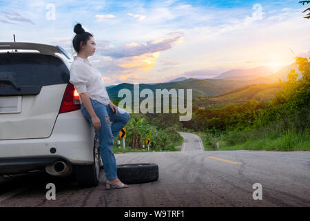 Junge schöne Frau in der Nähe von Auto für den Aufruf um Hilfe auf der öffentlichen Straße im Waldgebiet am Berg und Himmel Hintergrund Stockfoto
