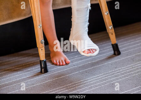 Gebrochenes Bein in Gips der unkenntlich kleines Kind. Junge Mädchen in der orthopädischen cast auf Krücken im Hause. Kind mit einem gebrochenen Bein auf Krücken, knöchelverletzung Stockfoto