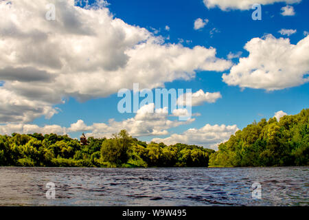 Blick vom Wasser aus auf den Fluss und einem bewaldeten Ufer, der Kegel der Dach eines hölzernen Turm im Wald vor einem blauen bewölkten Himmel, selektive Stockfoto