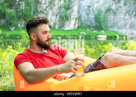 Freizeitaktivitäten Konzept, Camping, Sommerferien. Junge Bartgeier kaukasischen Mann mit Gläser Bier zur Festlegung von Orange Sofa. Entspannen und genießen das Leben auf der Luft b Stockfoto