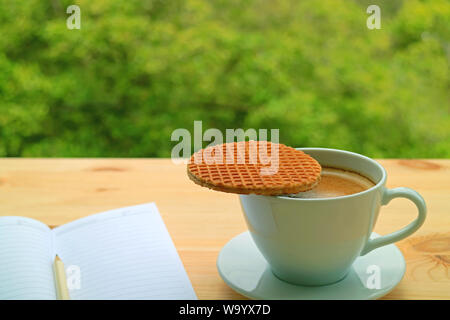 Heiße Tasse Kaffee mit einem stroopwafel auf seiner Spitze auf Fenster Seite Tabelle gegen Verschwommene grüne Laub serviert. Stockfoto