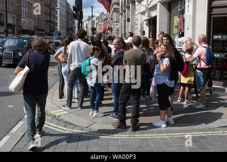 Eine Gruppe von spanischen Touristen versammeln, um ihre Führung zu hören, den Bürgersteig auf der Piccadilly blockieren und zwingt die anderen Fußgänger in auf-kommenden Verkehr, um sich, am 13. August 2019, in London, England. Stockfoto