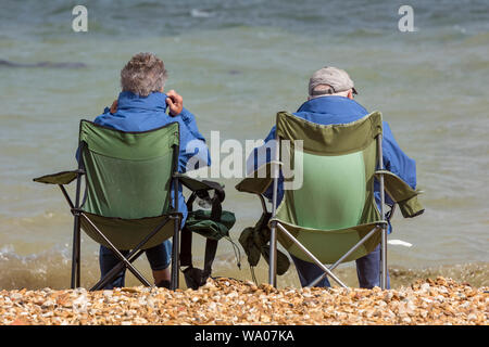 Ein älteres Rentner sitzt in zusammenklappbaren Stühlen am Strand am Meer und trägt Mäntel und Hüte. Stockfoto