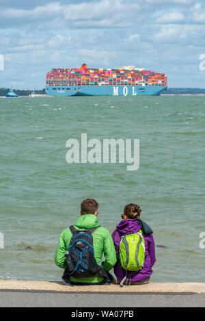 Ein junges Paar, das wasserdichte Jacken und Rucksäcke trägt, die auf einem Kieselstrand sitzen und auf ein großes Containerschiff in der solent at cowes blicken. Stockfoto