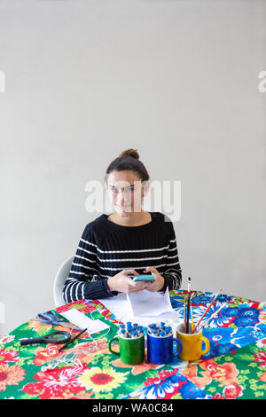 Junge talentierte Künstlerin eingeben Nachricht auf Handy am Tisch sitzen mit blühenden Handtuch in Ihrem Studio mit weißem Hintergrund. Verwendet die Stifte, Papier, Stockfoto