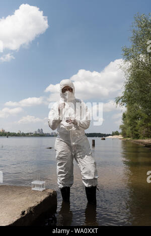 Volle Länge Blick auf Wasser Inspektor im schützenden Kostüm, Latex Handschuhe und Atemschutzmaske holding Kolben mit Wasser Probe am Fluss Stockfoto