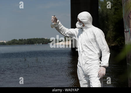 Wasser Inspektor im schützenden Kostüm holding Kolben mit Wasser Probe am Fluss Stockfoto