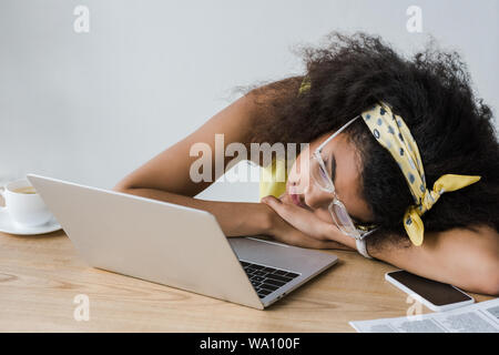 Afrikanische amerikanische Mädchen liegt auf dem Tisch in der Nähe der Schale mit Getränk und Laptop müde Stockfoto