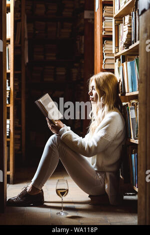 Seitenansicht der blonden Frau mit Buch und das Sitzen auf dem Boden in der Bibliothek Stockfoto