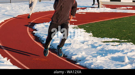 Jungen laufen schnell in der Spur einer roten Piste mit Schnee auf beiden Seiten im Winter Leichtathletik Praxis. Stockfoto