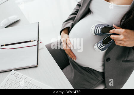 7/8-Ansicht der schwangeren Frau mit kleinen Baby Stiefeletten beim Sitzen hinter Tabelle mit Tastatur und Notebook Stockfoto