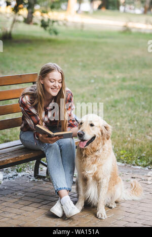 Volle Länge Blick auf schöne junge Mädchen in Freizeitkleidung lesen Buch und einen Streichelzoo golden retriever während auf Holzbank im Park sitzen Stockfoto