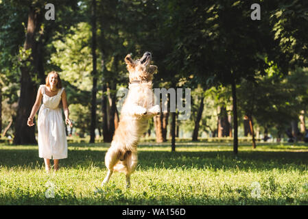 In voller Länge ansehen der jungen Mädchen in weißem Kleid lächelnd und Suchen im jumping Golden Retriever auf Wiese Stockfoto