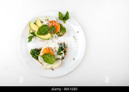 Spiegeleier auf einem Toast Brot mit Avocado, Spinat und Samen auf einem weißen Teller auf dem weißen Hintergrund Stockfoto