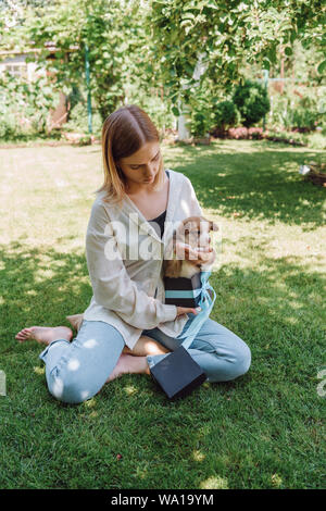 Blonde Mädchen barfuß in den grünen Garten mit niedlichen Welpen in Geschenkbox Stockfoto