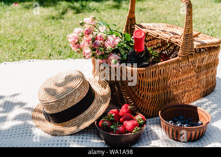 Weidenkorb mit Rosen und eine Flasche Wein auf weiße Decke in der Nähe von Stroh Hut und Beeren an sonnigen Tag im Garten Stockfoto